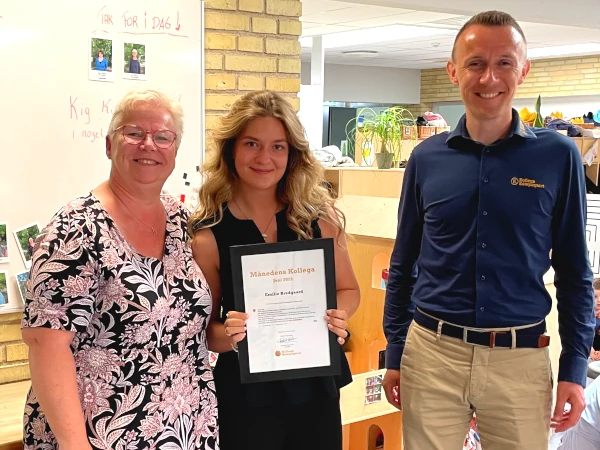 Månedens Kollega i juni 2025 er Emilie Bredgaard (i midten) fra Børnehuset Nordlys i Vordingborg Kommune. Mette Boye (t.v.) havde nomineret Emilie, som fik en gavekurv og et diplom. På billedet ses også Frederik Karstrøm, direktør i KollegaKompagniet.