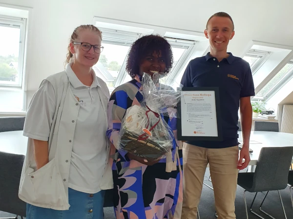 Månedens Kollega i august 2025 er Josie Ingabire (i midten) fra Omsorg Sjælland. Susanne Bay Breiting (t.v.) havde nomineret Josie, som fik en gavekurv og et diplom. På billedet ses også Frederik Karstrøm, direktør i KollegaKompagniet.
